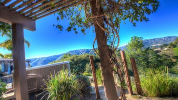 A wooden pergola with a bench, surrounded by lush greenery and a mountainous backdrop.