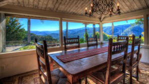 A dining area with a wooden table and chairs, large windows offering a view of mountains and greenery. - Lx16: Valley View Estate Above The Wineries With Hot Tub (Carmel Valley)