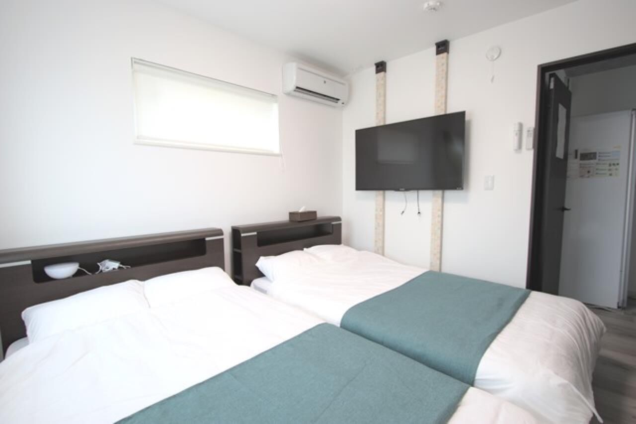 Een hotelkamer met twee bedden, een aan de muur bevestigde televisie, een airconditioning en een raam met jaloezieën.