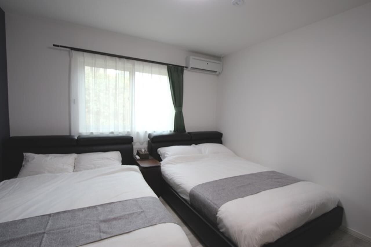Hotellihuone, jossa on kaksi erillistä sänkyä, ikkuna, jossa on läpikuultavat verhot, ilmastointilaite ja yöpöytä, jossa on lamppu.