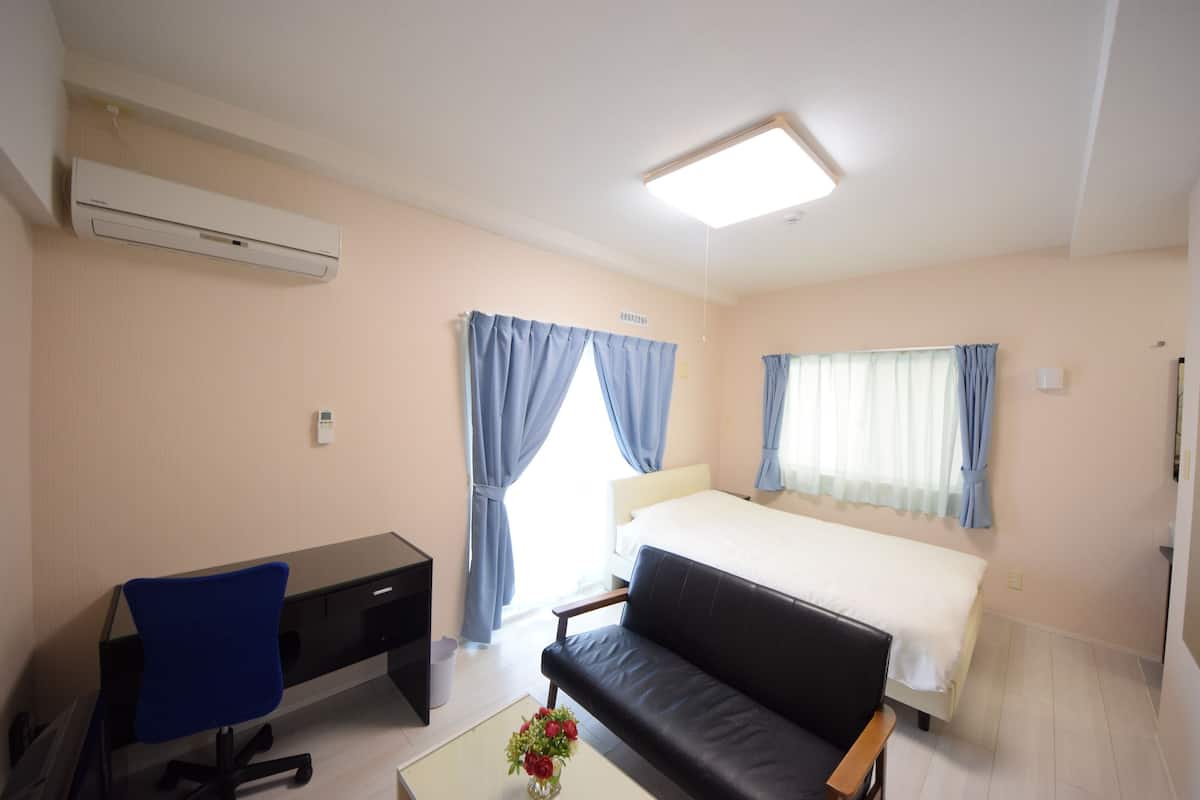 A hotel room with a bed, a desk with a chair, a sofa, a coffee table with flowers, and an air conditioning unit.