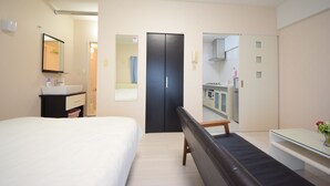 A hotel room with a bed, a small desk, a bench, a mirror, a door, and a kitchenette. - SUMUKA Shintoshin II (Naha)