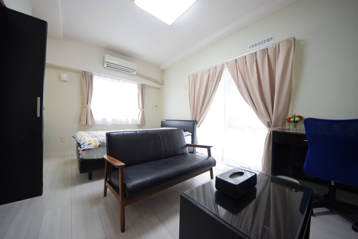 A hotel room with a black leather sofa, a glass coffee table, a desk with a chair, a bed with a colorful blanket, and large windows with curtains.