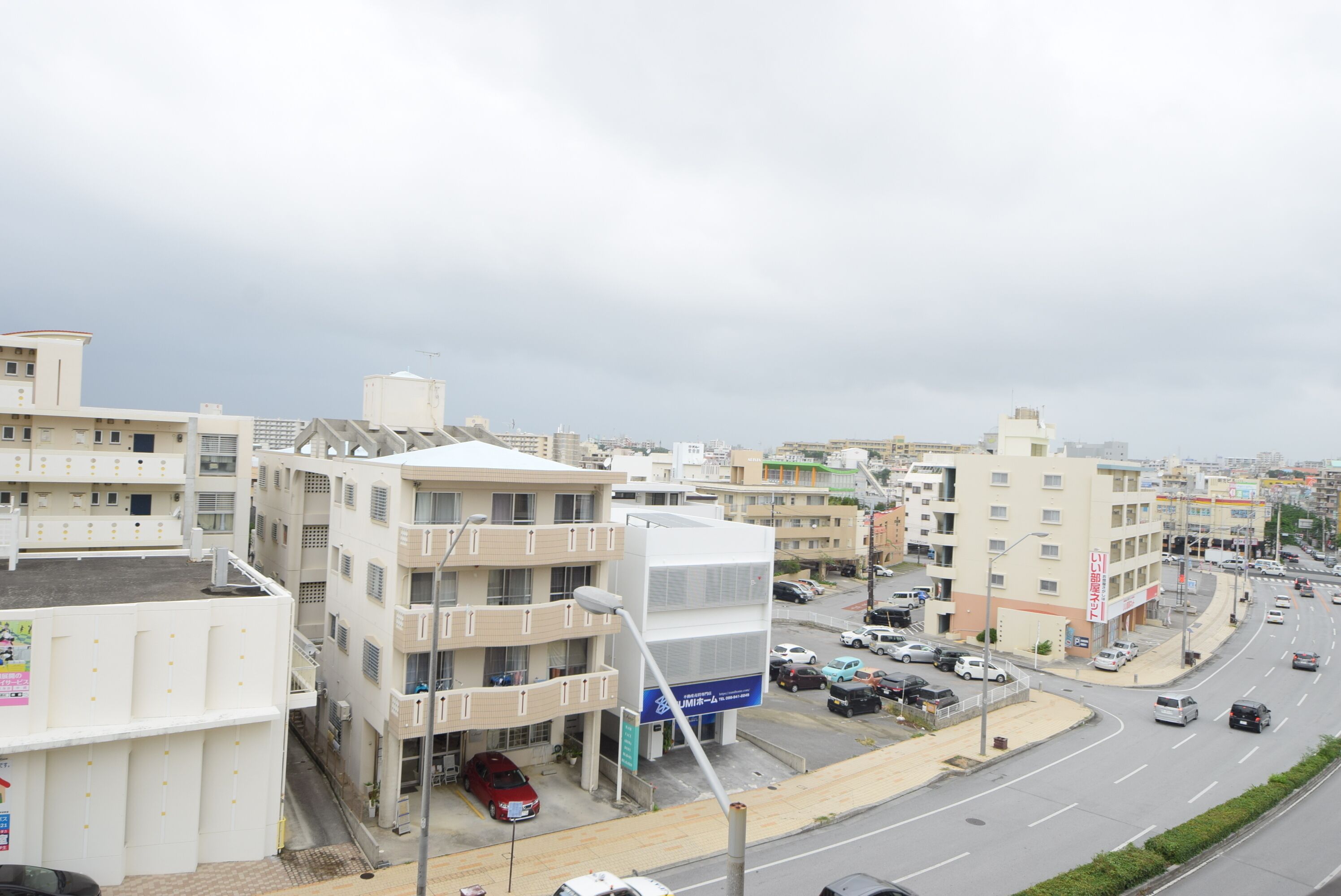 A multi-story building with a flat roof, several balconies, and a parking area with cars.