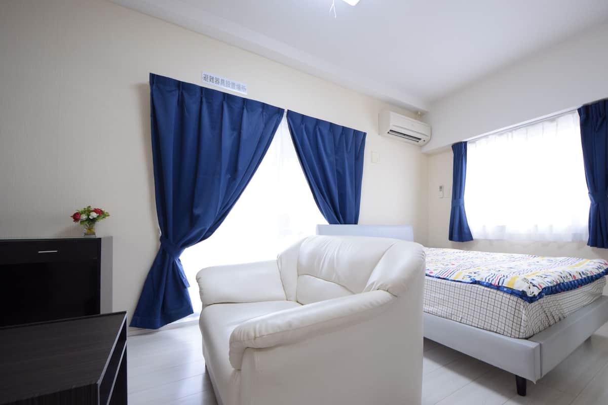 A bedroom with a bed, a white armchair, a desk, and a window with blue curtains.