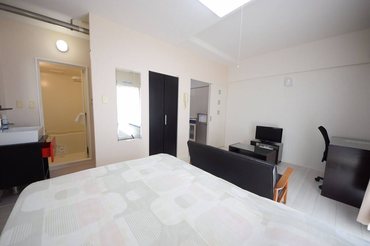 A hotel room with a bed, a desk, a chair, a TV, and a bathroom with a shower and toilet.
