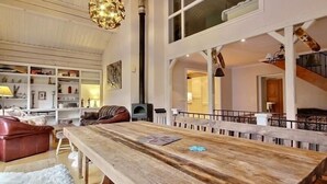 Een ruime woonkamer met een grote houten eettafel, een open haard en een hoge plafon met zichtbare balken.