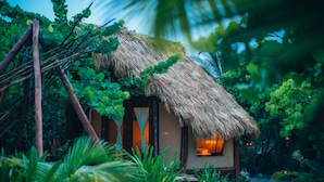 A thatched-roof hut with a bed visible through the window, surrounded by lush greenery and a sandy ground. - Glamp Ikal Tulum (Tulum)