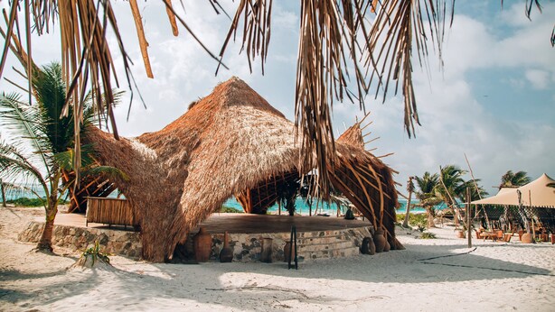 A thatched-roof structure with a stone base, surrounded by palm trees and a sandy beach. - Glamp Ikal Tulum (Tulum)