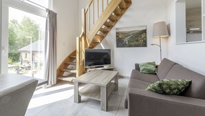 Ein modernes Wohnzimmer mit einer Holztreppe, einem Flachbildfernseher, einer grünen Sofaausstattung und einem großen Fenster mit Blick ins Freie.