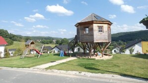 Ein Spielhäuschen aus Holz auf Stelzen mit rotem Dach, umgeben von einer Rasenfläche und einem Spielplatz.