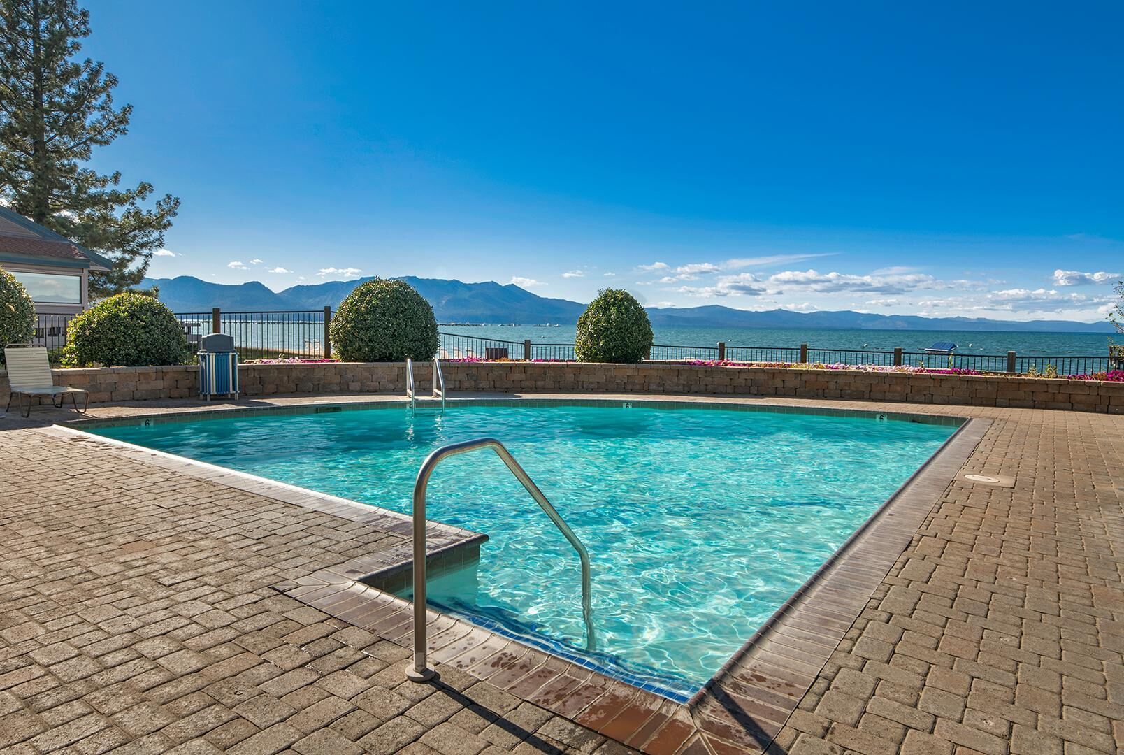 A rectangular swimming pool with clear blue water, surrounded by a brick deck and a metal railing. In the background, there is a view of mountains and a body of water.