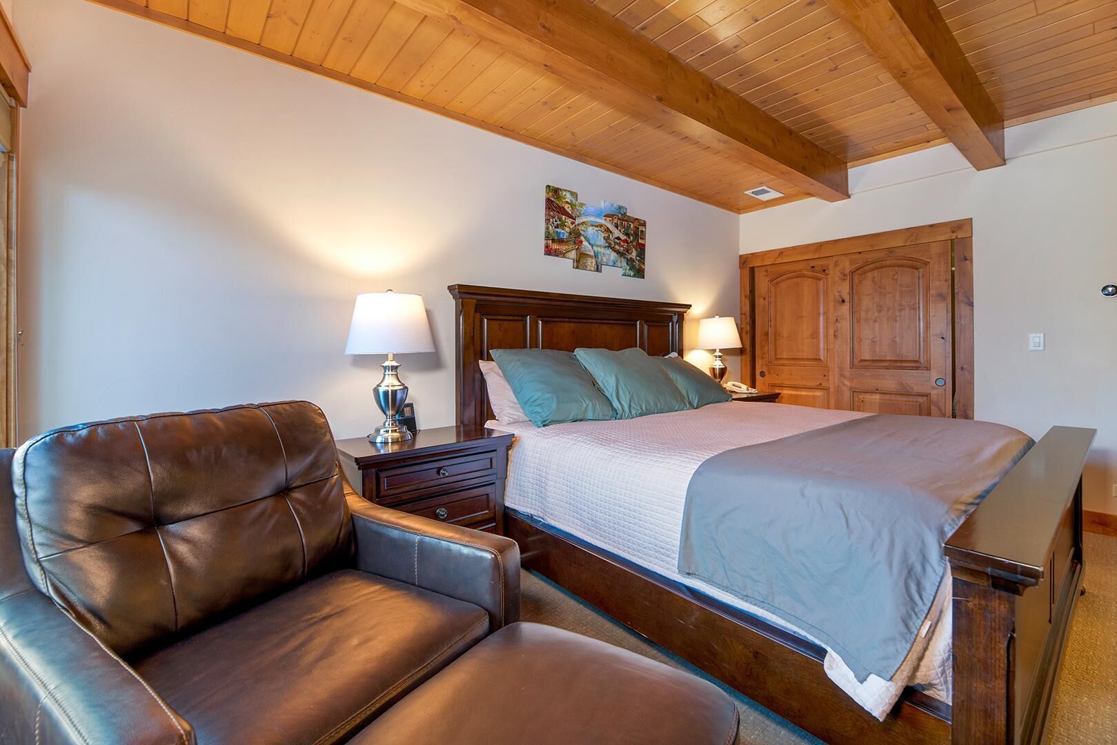 A bedroom with a wooden ceiling, a bed with a headboard, a nightstand with a lamp, a leather armchair, and a wooden door.