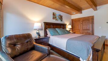 A bedroom with a wooden ceiling, a bed with a headboard, a nightstand with a lamp, a leather armchair, and a wooden door.