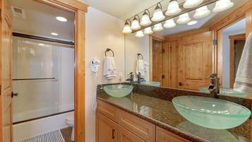 A bathroom with a glass bowl sink, wooden cabinets, a large mirror, and a shower.