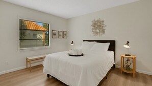 A neatly arranged bedroom with a large bed, a bench, a bedside table with a lamp, and a wall-mounted abstract art piece.