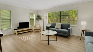 A living room with a flat-screen TV, a grey sofa, a round coffee table, and large windows with blinds.