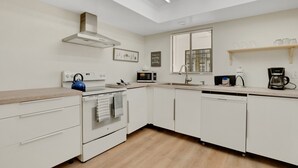 A modern kitchen with white cabinetry, a built-in oven, microwave, and dishwasher. There is a window with blinds, a shelf with glasses, and a kettle on the counter.