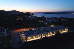 A modern building with a flat roof, surrounded by a parking area and a body of water in the background. - Villa Lana - Home4You (Kraj)