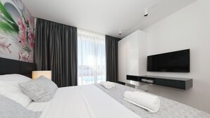 A modern hotel room with a large bed, a flat-screen TV, a desk with a lamp, and a view of the outdoors. - Homes4You Villas Complex (Kraj)