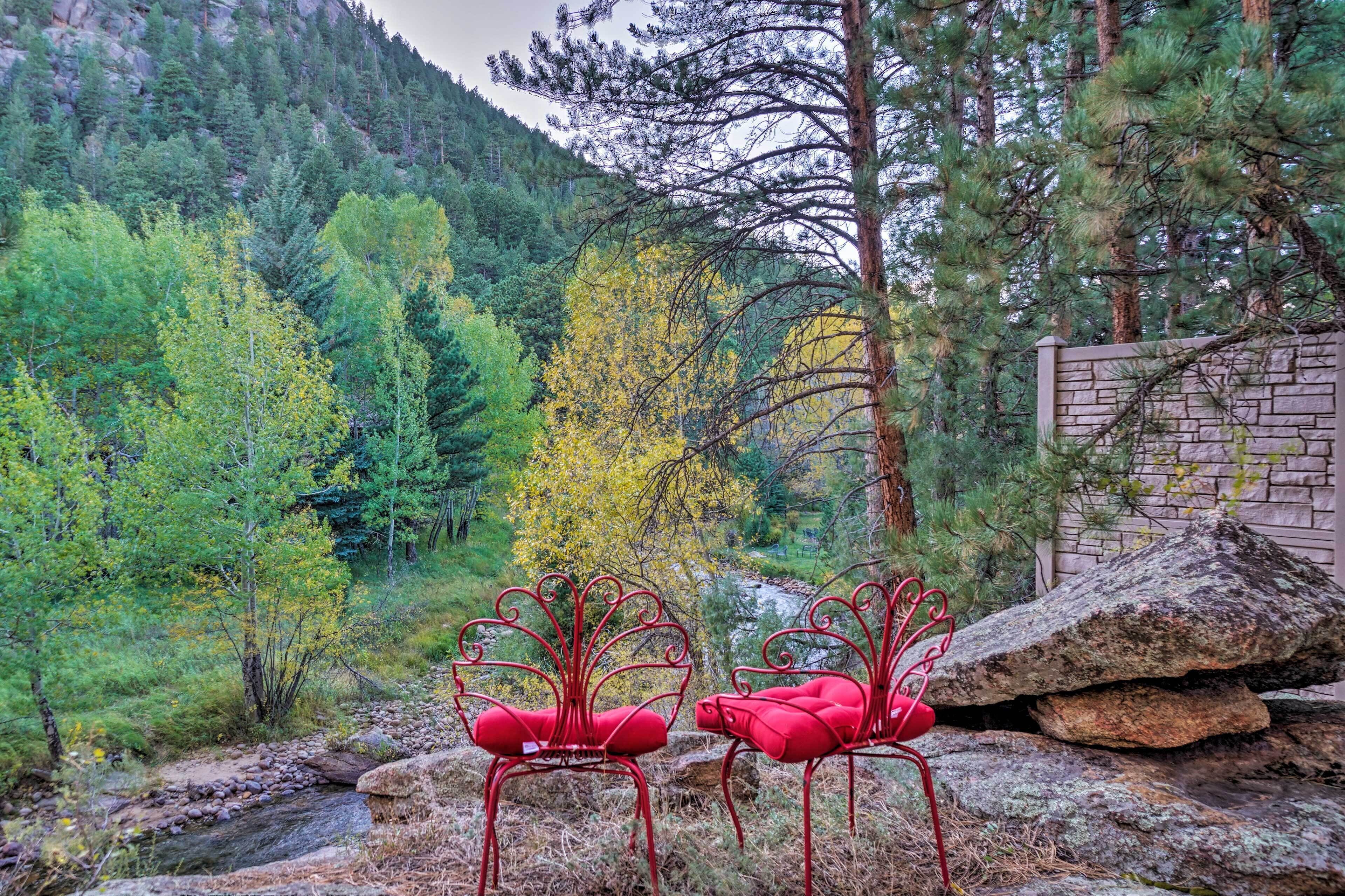 Two red metal chairs with intricate designs, positioned on a rocky surface with a forested mountain backdrop.