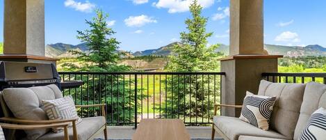 A balcony with a sofa, a table, and a grill, overlooking a mountainous landscape.