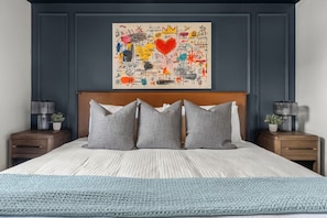 Une chambre à coucher avec un lit, des oreillers gris, une tête de lit bleue, une œuvre d’art murale et des tables de chevet avec des lampes.