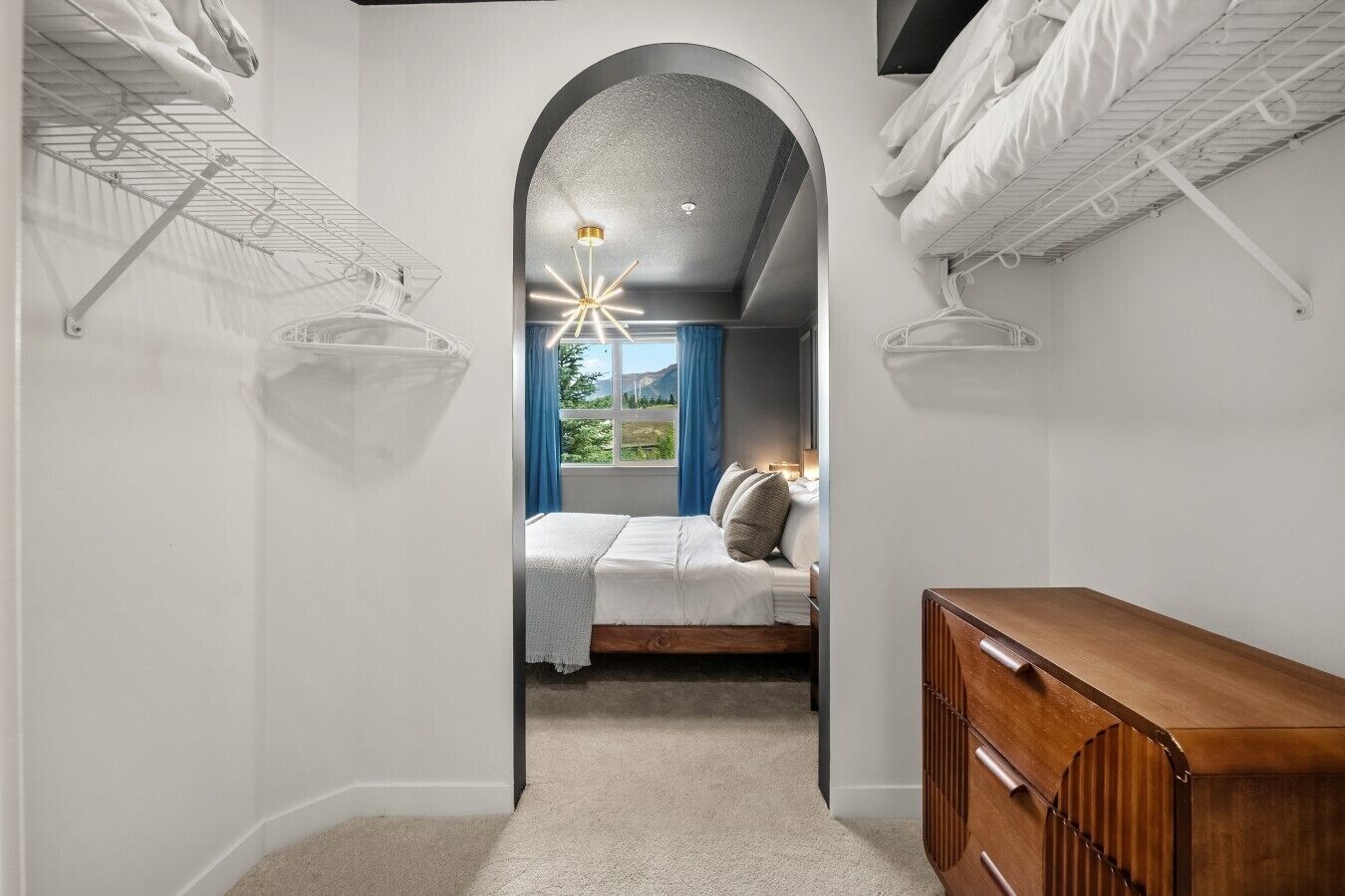 Une chambre à coucher avec un lit, une commode en bois, une fenêtre avec des rideaux et un couloir menant à une autre pièce.