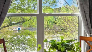 A room with a large window offering a view of a lake, a dock, and a small building. There is a wooden chair and a table with a potted plant in the foreground. - Lake Anna Waterfront - Sleeps 21 - Dock for 3 Boats (Orange)