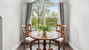 A dining area with a round table, two wooden chairs, a potted plant, and a view of trees and a building through a large window. - Lake Anna Waterfront - Sleeps 21 - Dock for 3 Boats (Orange)