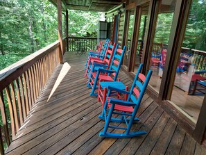 Une terrasse en bois, agrémentée de fauteuils à bascule et de tables, entourée d’arbres.