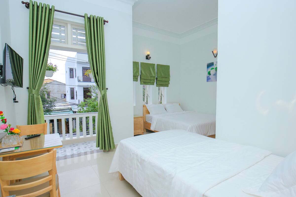 A hotel room with two beds, a balcony with green curtains, a small table with a chair, and a TV mounted on the wall.