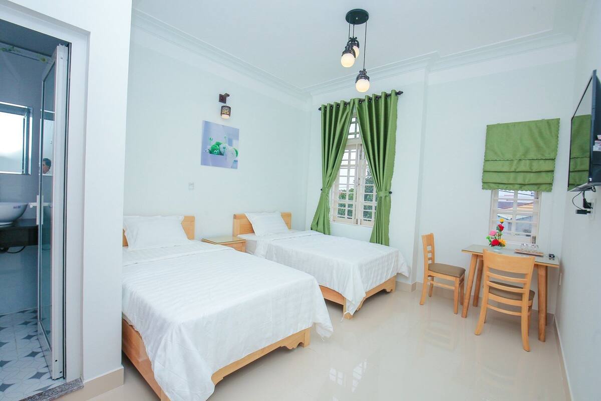 A room with two beds, a desk, and a chair. There is a window with green curtains and a picture on the wall.