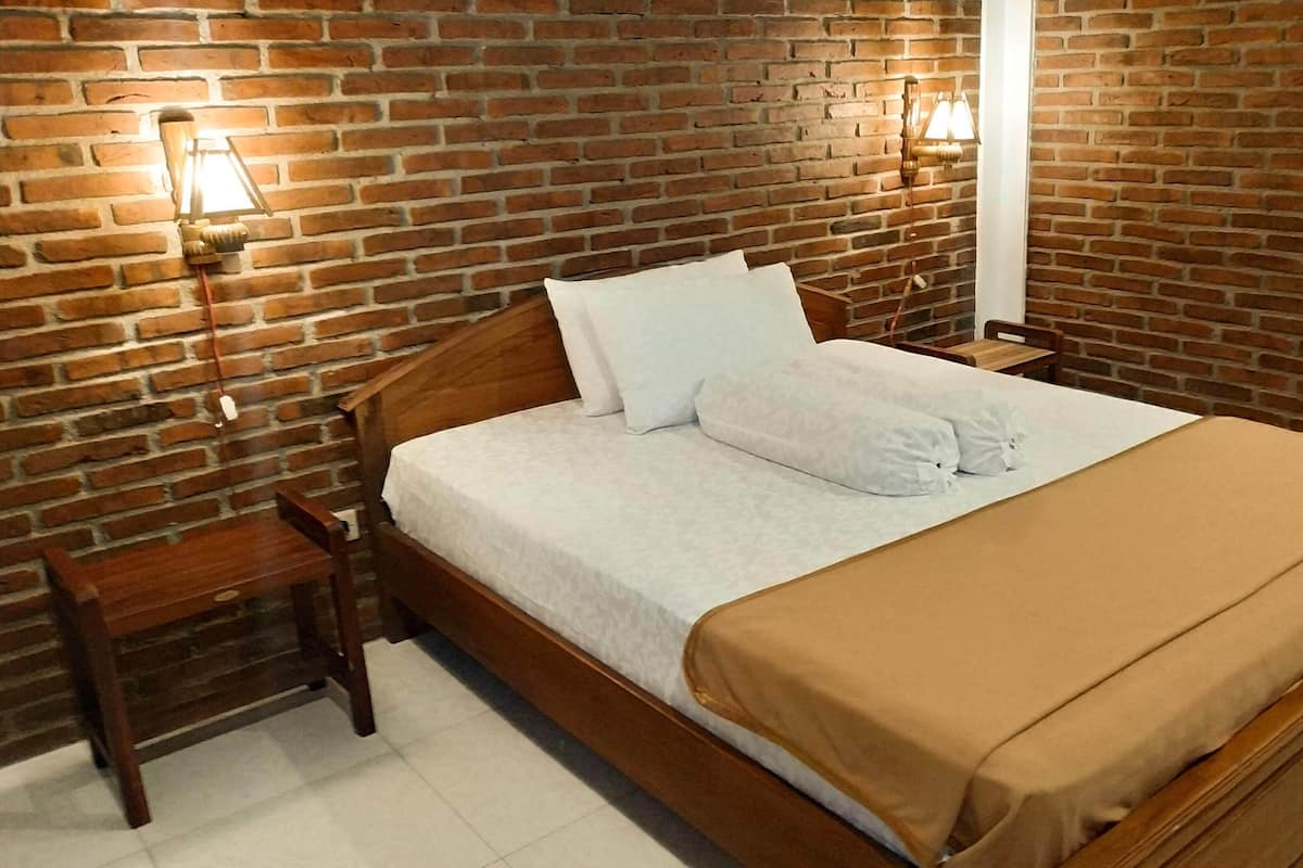 A neatly made bed with a wooden headboard, white linens, and a brown throw blanket. There are two bedside tables with lamps, and a brick wall in the background.