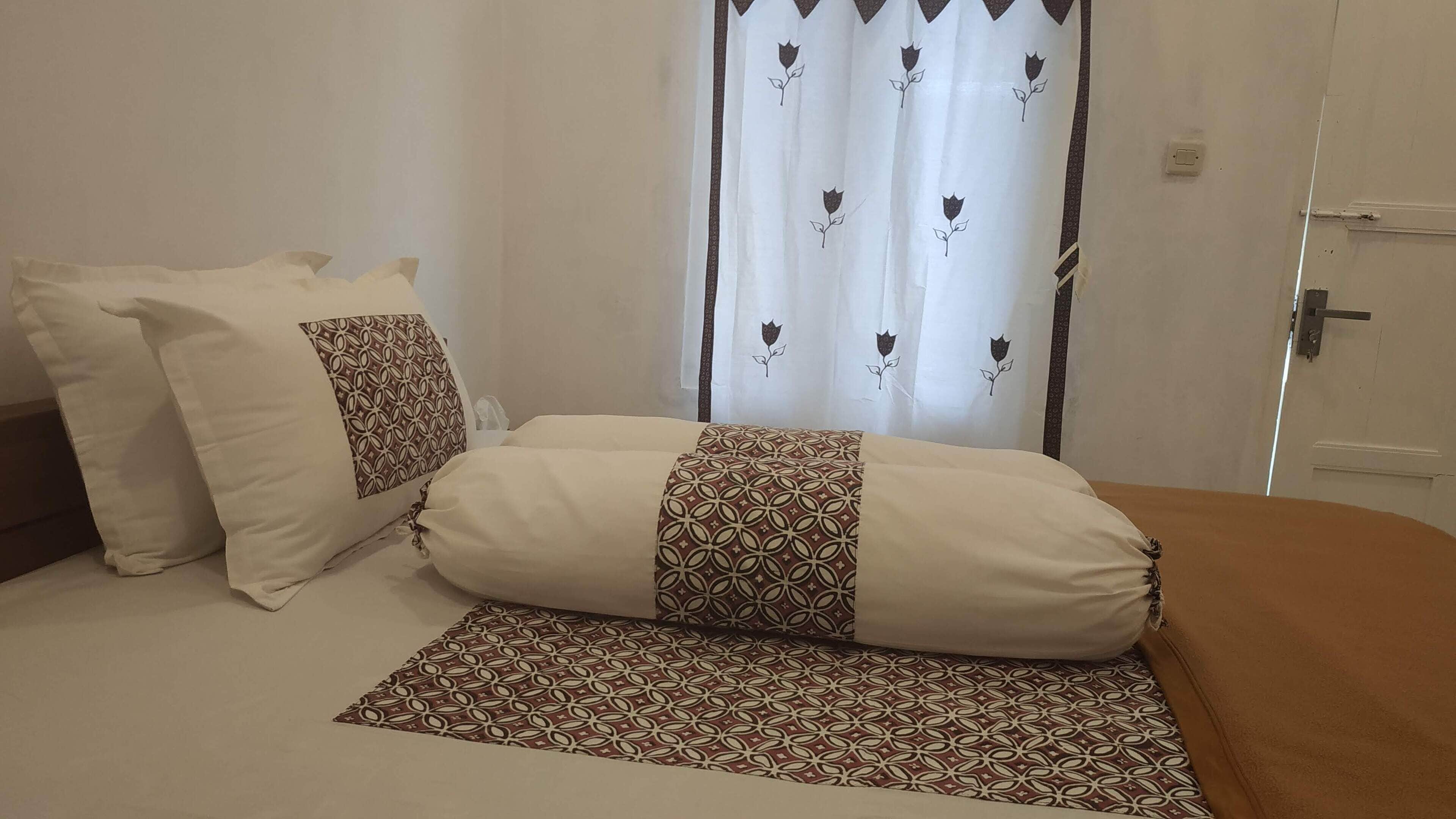 Un lit bien fait, agrémenté d’oreillers à motifs et d’un couvre-lit assorti, un rideau blanc orné de motifs floraux et une porte dotée d’une simple poignée.