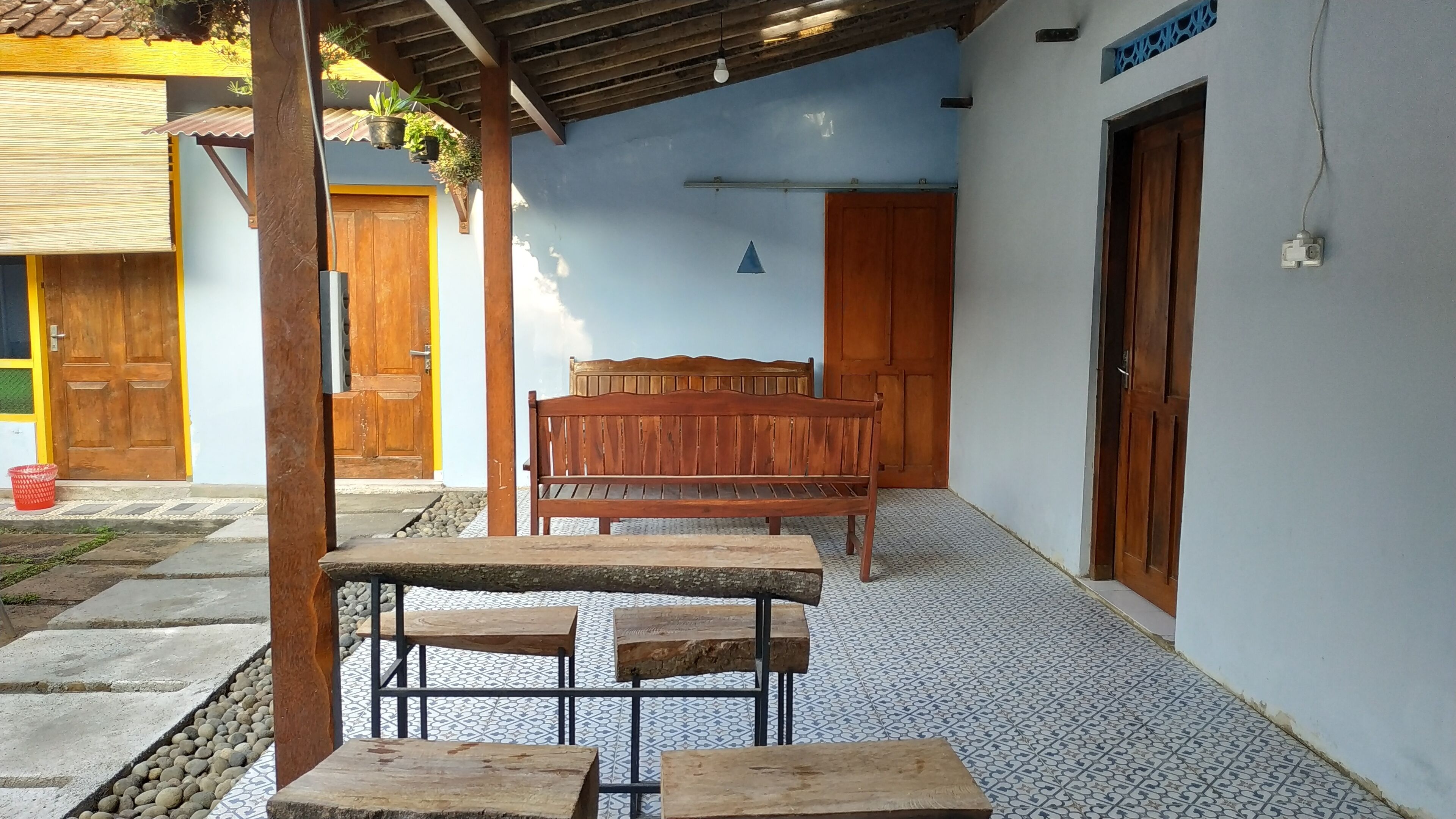 A covered outdoor seating area with a wooden bench and tables, a tiled floor, and a blue wall with a wooden door.