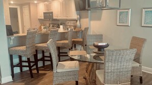 A dining area with wicker chairs, a glass table, and a modern kitchen in the background.