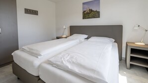 Una cama doble con sábanas blancas, una mesita de noche con lámpara, un interruptor de luz en la pared y un cuadro con un paisaje.