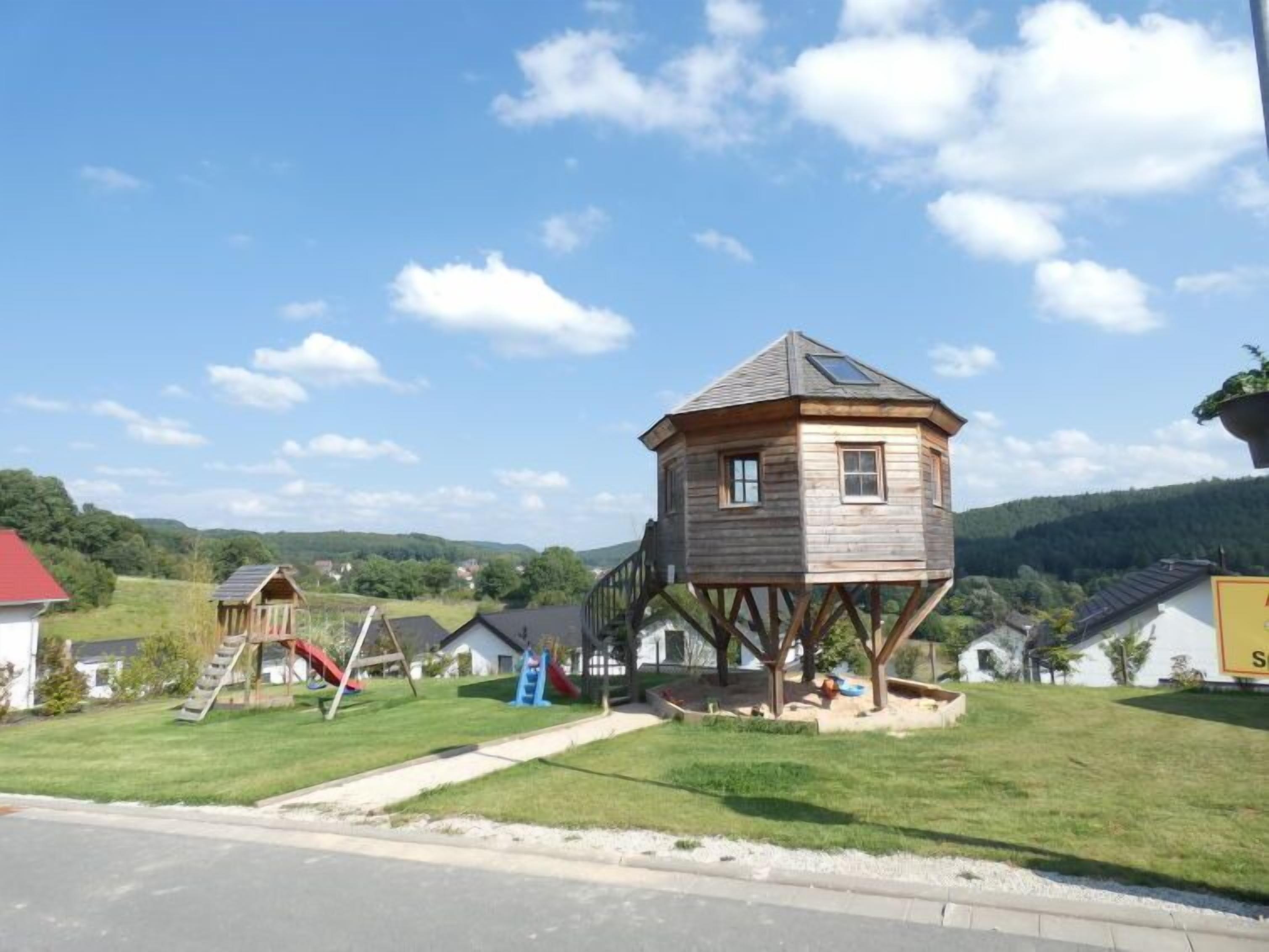 A wooden playhouse on stilts with a red roof, surrounded by a grassy area and a playground.
