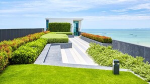 A modern building with a glass door, surrounded by well-maintained hedges and a grassy area leading to a balcony overlooking the sea. - Pony's Homestay -The Base Condo- (Bang Lamung)