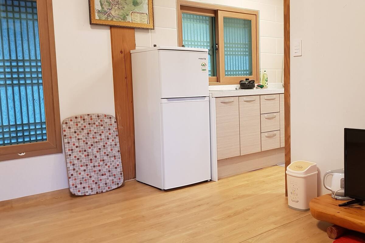 Une cuisine compacte équipée d’un réfrigérateur, d’un four à micro-ondes et d’une machine à café. On y trouve un meuble en bois avec des tiroirs et une petite poubelle. Un téléviseur est posé sur un meuble en bois.