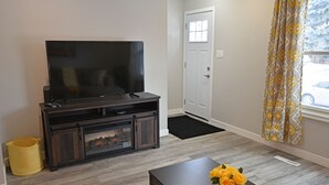Uma sala de estar com televisão sobre um suporte de madeira, uma porta e uma janela com cortinas.