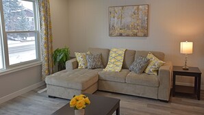 Uma sala de estar com um sofá seccional bege, almofadas estampadas, uma mesinha com um vaso de flores amarelas, um quadro na parede e uma janela com cortinas.