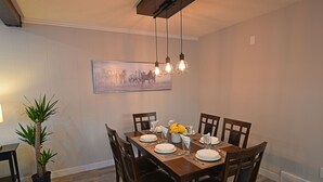 Uma área de refeições com uma mesa de madeira posta para quatro pessoas, rodeada por cadeiras. Há uma planta em vaso à esquerda e um quadro na parede acima da mesa.