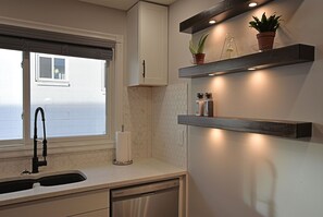 A kitchen with a window, a sink, a dish rack, a potted plant, and a shelf with kitchen items. - EXQUISITE 3BD HOUSE CLOSE TO AIRPORT, PL HOSP, DOWNTOWN, SLEEPS 7 (Calgary)