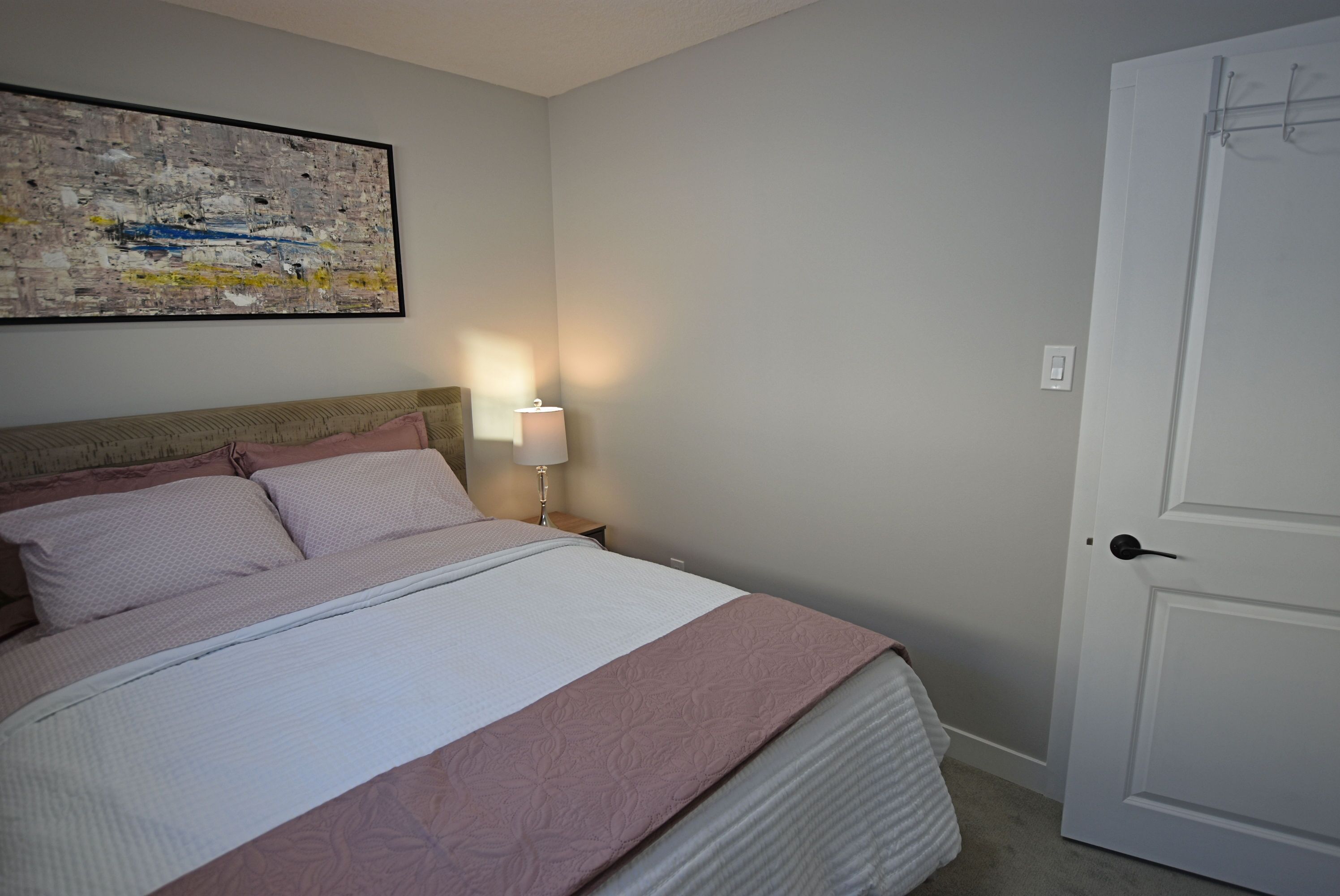 A neatly made bed with a pink and white bedspread, a nightstand with a lamp, and a door to the right.