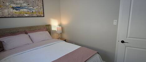A neatly made bed with a pink and white bedspread, a nightstand with a lamp, and a door to the right.