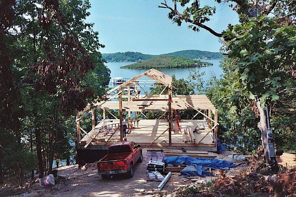 Une cabane en construction, située près d’un lac, d’où l’on a une vue dégagée sur l’eau et les collines en arrière-plan.