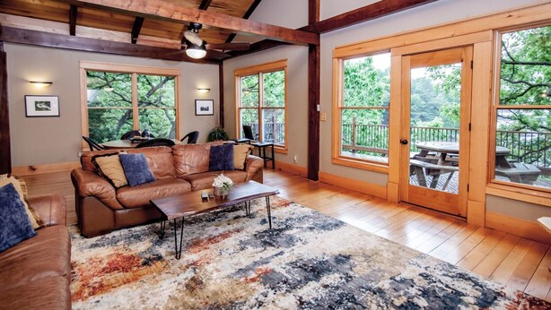 A cozy living room with a leather sofa, a coffee table, and a large rug. There are two windows with a view of trees and a balcony with a table and chairs. - Free Night: Make Ozarks Memories at this Custom Lakefront Post & Beam Home VIDEO (Eldon)