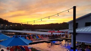 A poolside area with lounge chairs and umbrellas, a pool, and string lights against a sunset sky. - Free Night: Make Ozarks Memories at this Custom Lakefront Post & Beam Home VIDEO (Eldon)
